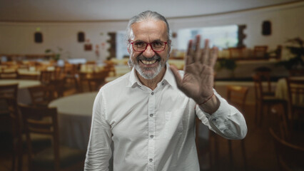 Fototapeta premium Man waves hand inside restaurant building, smiling and showing open palm while wearing white shirt and red glasses; friendliness welcome.