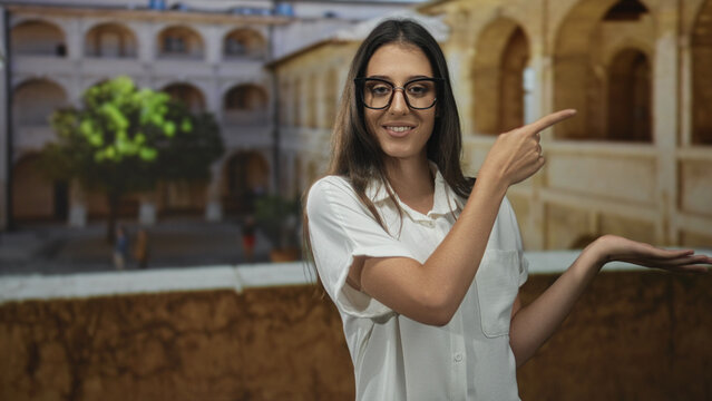 Woman points fingers to the left on an old town street, wearing glasses and white shirt, bare forearms visible; skepticism doubt indecision.