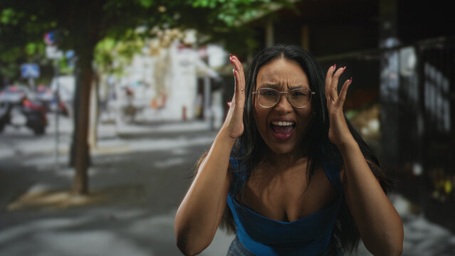 Woman with hands at her head and mouth open showing tongue, wearing blue top and glasses, reaching toward camera on a street; exasperation moment.