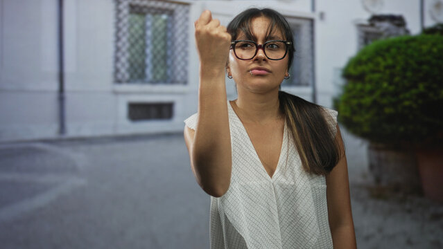 Woman raising hand in a shrug gesture on a street in front of a building, glasses and white blouse visible; indifference casual attitude.