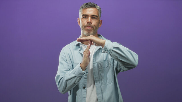 Man middle age grey hair making time out gesture with hands in studio purple backdrop wearing light denim shirt and white tee; boundary assertion.