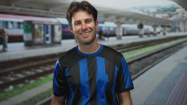 Man in blue soccer jersey smiling at camera on train platform inside a station building near railway tracks; contentment travel.