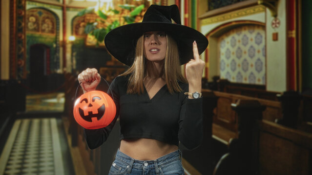 Young woman in witch hat holding pumpkin bucket, showing middle finger and bare midriff inside ornate church; rebellious defiance.