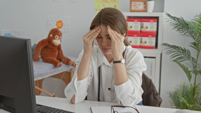 Young woman doctor with stethoscope presses hands to forehead at pediatric clinic desk; fatigue concern.