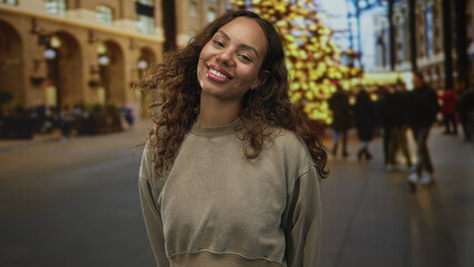 Fototapeta premium Woman smiles with visible face and slight head tilt on busy city street beside lit holiday tree and blurred crowd; joy confidence.