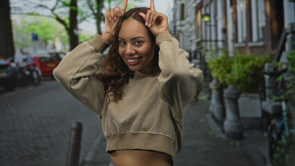 Fototapeta premium Woman showing midriff, smiling and holding fingers as horns on a downtown street; playful carefree joy.