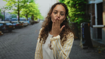 Fototapeta premium Woman smiling with visible face and curly hair on cobblestone street in city wearing white tee and striped shirt looking at camera; joy.