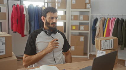 Fototapeta premium Man adjusting headset while seated by stacked shipping boxes and clothing racks in a building; frustration.