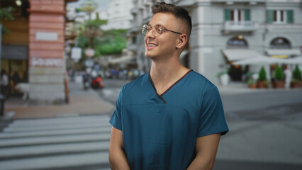 Fototapeta premium Young doctor in teal scrubs smiles, hands clasped at waist and head tilted while standing on a crosswalk in an urban street with cafe terraces; optimism.