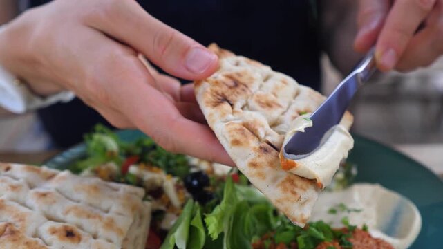 Person spreading creamy hummus dip on a piece of fresh pita bread with a knife, ready to eat a healthy mediterranean meal with various salads and appetizers on a green plate