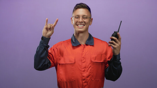 Young mechanic man wearing orange coverall and glasses, smiling and holding a walkie talkie while flashing rock hand sign in studio; playful joy.