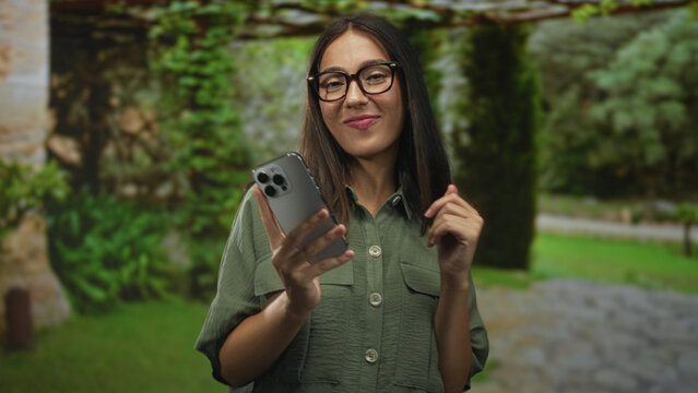 Young hispanic woman with glasses and brunette hair smiling while holding smartphone and typing with hands in a forest park with green foliage and stone path; contentment.