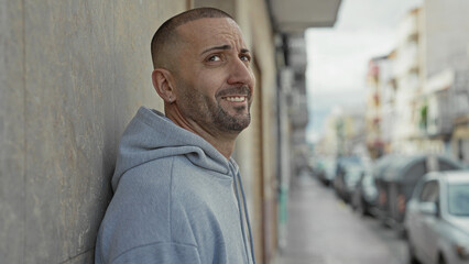 Man leaning against wall, ear visible, smiling with hood up and light stubble, looking up on street  quiet contentment. © Krakenimages.com