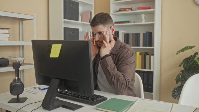 Young man at computer rubs forehead and pinches nose while seated at desk in studio; stress concentration overload.