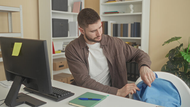Young man peels banana at keyboard while typing at computer at desk in building with bookshelf and backpack visible; focus snack break.