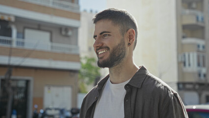 Man smiling showing earring and beard, wearing a casual shirt, standing on a sunlit street by an...