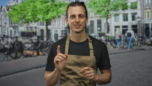 Young man with hands raised palms out on street in apron and black shirt, long hair tied back; apology humility.