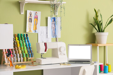 Tailor's workplace with blank laptop and sewing machine near green wall in atelier