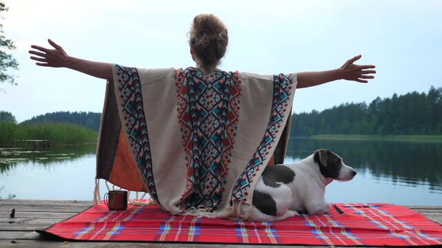 Serene woman in a traditional poncho meditating with her dog on a wooden pier, she raises her arms embracing the tranquil lake and forest landscape, feeling connected with nature