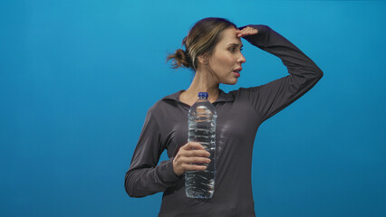 Obraz premium Woman holding a large clear plastic water bottle and shielding her eyes with one hand in blue studio backdrop, angled profile, sweaty and panting; thirst.