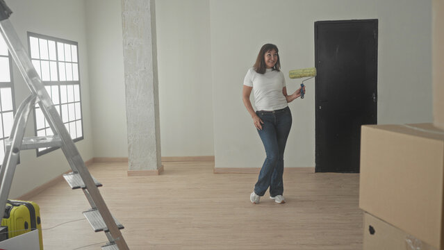 Woman with paint roller holds roller in right hand, hand on hip, standing in empty building room with boxes and ladder; home renovation hopeful.