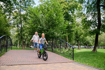Fototapeta premium Family with children riding bicycles and playing in green park, active outdoor lifestyle. Multi-generational family spending time together in a lush green park. Grandmother walking with two young boys