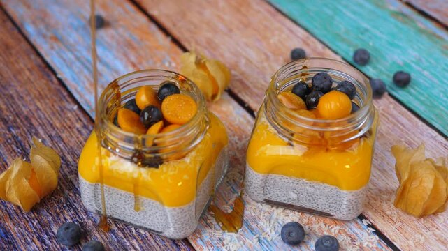 Delicious honey being poured onto two glass jars of fresh chia seed pudding layered with mango mousse and topped with fresh blueberries and physalis berries on a rustic wooden table