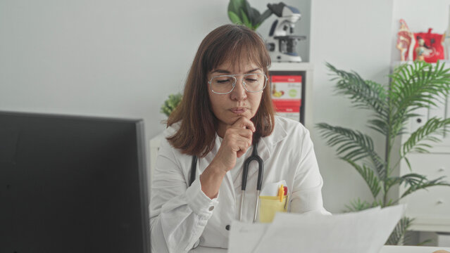 Woman doctor reading papers at computer desk with stethoscope in clinic building; patient care concern.