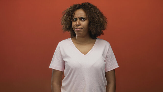 Woman furrows brow with visible face and curly hair wearing pale pink t shirt in orange studio; confusion doubt.