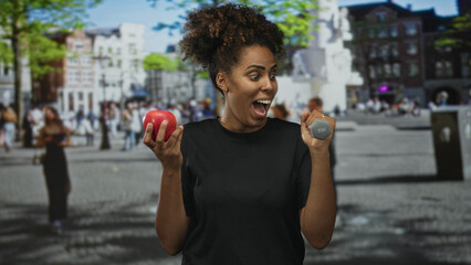 Fototapeta premium Woman holding apple and dumbbell while smiling on busy street plaza with buildings and trees visible; healthy choice joy.