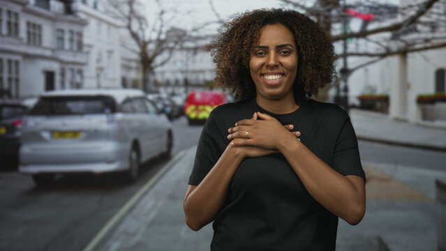Woman clasping hands over chest on a city street lined with parked cars and white townhouses; gratitude.