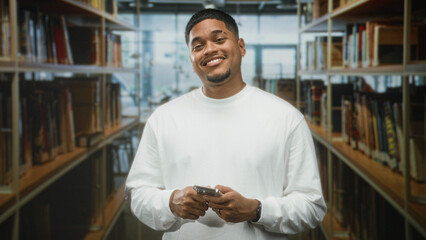 Man with beard in white shirt holding smartphone with both hands and smiling while typing between bookshelves in library building  relaxed contentment. © Krakenimages.com