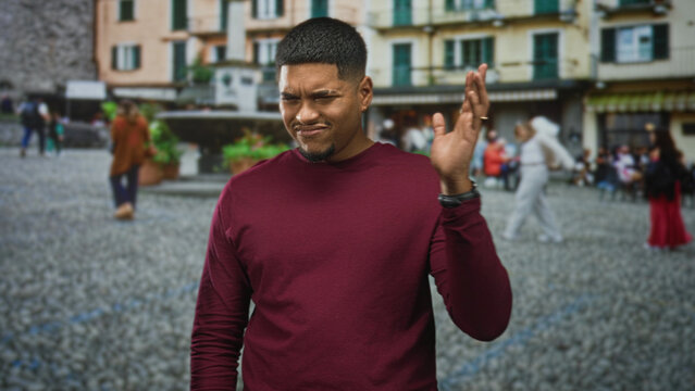 Man with beard and crewcut waving hands and grimacing on a cobblestone street in front of buildings and fountain; disgust.