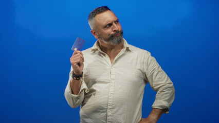 Fototapeta premium Man holding creditcard up with hand on hip and bracelets while posing in blue studio wall; confidence money choice.