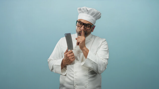 Man chef middle aged with grey hair holding cleaver and finger to lips for silence in studio with light blue backdrop; mischief.