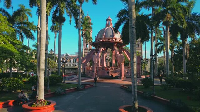 Tampico, Tamaulipas Mexico; February 2 2026. Pink Octopus Kiosk and Tampico Cathedral are shown in a low angle aerial drone dolly in passing between palm trees under a clear blue sky.