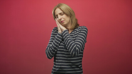 Fototapeta premium Woman with hands to cheek and closed eyes making sleeping gesture, wearing striped shirt against red studio backdrop; tired rest sleepiness.