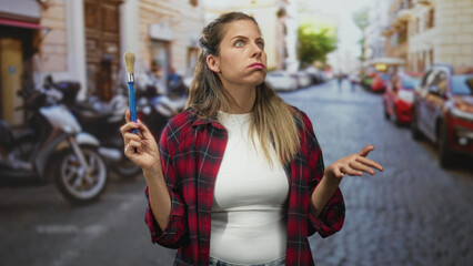 Fototapeta premium Woman painter holds brush up in hand and looks upward on street, wearing red plaid shirt and white top; creative contemplation.