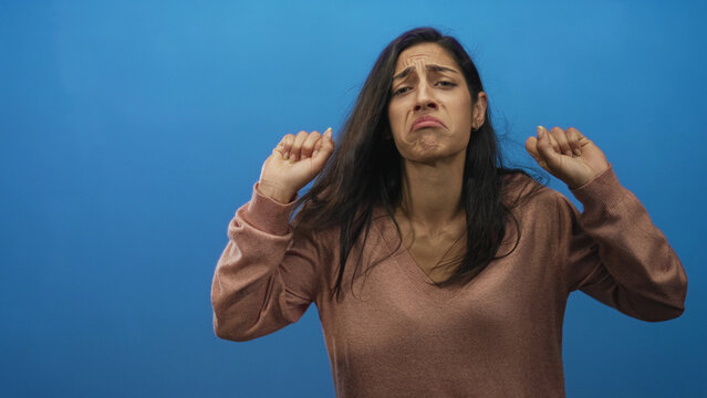 Woman wearing brown vneck sweater making a grimacing tongue out face with clenched fists and closed eyes in blue studio; disgust.