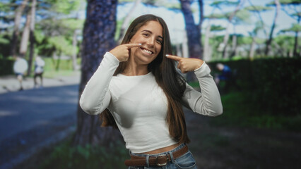 Fototapeta premium Young woman pointing fingers to cheeks and smiling while wearing a shirt and belt in a forest path with trees and runners visible; confidence joy.