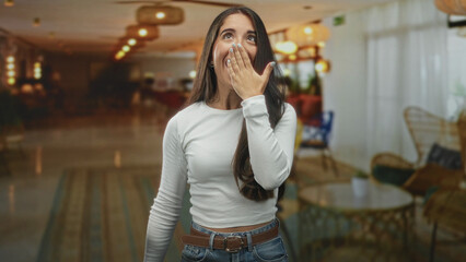 Fototapeta premium Young hispanic woman with hand over mouth smiling in a hotel lobby building wearing white top and jeans, casual pose; playful joy.