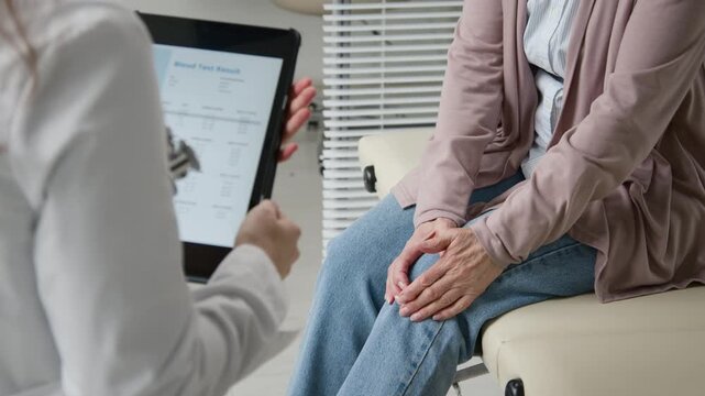 Rack focus shot of female doctor holding digital tablet with blood test results displaying on screen while treating elderly woman for knee pain in examination room