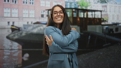 Obraz premium Woman hugging arms, smiling, wearing glasses and blue coat in front of canal boat and building; smiling contentment.