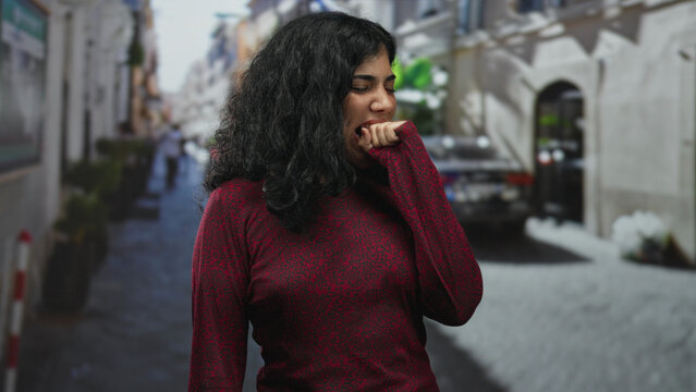 Young brunette woman in red sweater bites her fist on a narrow city street, hand partly in mouth while suppressing a yawn and closing eyes; tired solitude.