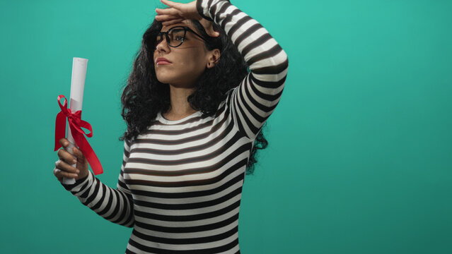 Young brunette woman holding a rolled diploma tied with red ribbon and hand to forehead shielding eyes in a teal studio; achievement pride.