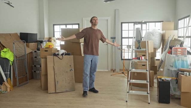 Man arms outstretched amid stacked cardboard boxes, ladder, tools and bubblewrap in a studio; fresh start relief.