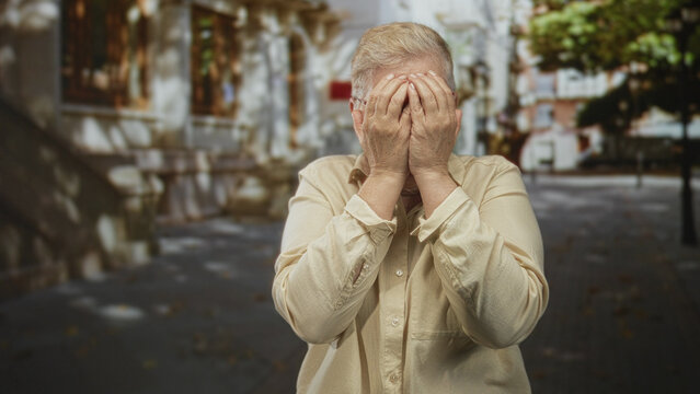 Middle aged woman with hands on cheeks and tongue out on city street wearing glasses and beige shirt, playful pose; silliness spontaneity joy.