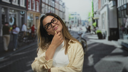 Obraz premium Woman touching chin with hand on a city street wearing glasses and casual shirt; contemplation quiet curiosity.