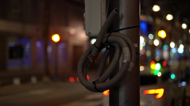 Close up of electrical cables attached to a pole at night with blurred city lights and traffic in the background.