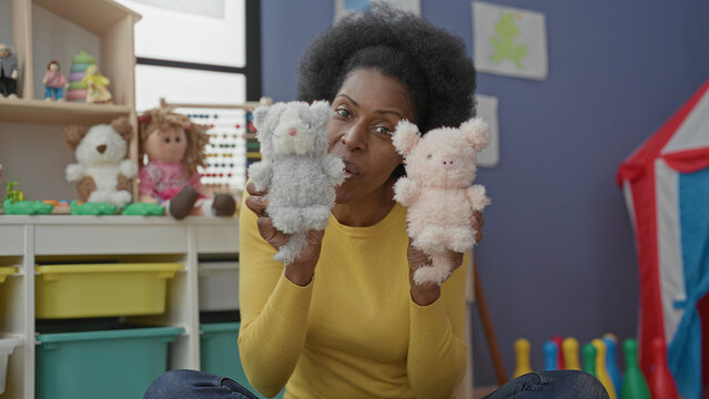 Woman holding two plush hand puppets while speaking in a colorful preschool classroom; playful learning.